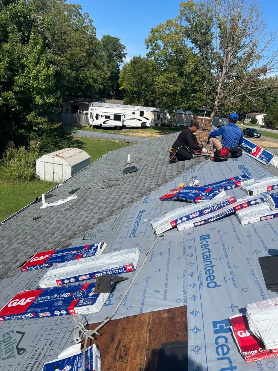 Roofing installers working near a chimney with shingles, underlayment and staged bundles visible